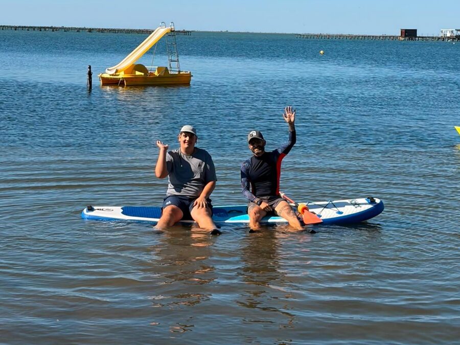 Usuari d'APASA i monitor asseguts sobre una taula de paddle surf a la platja